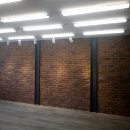 Wnętrze z ceglaną ścianą, wzmocnioną pionowymi, czarnymi profilami stalowymi, oświetlone rzędem lamp jarzeniowych pod sufitem. Podłoga wyłożona panelami imitującymi drewno.