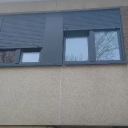 Szare okna z roletami zewnętrznymi zamontowane w budynku z elewacją z szarego tynku strukturalnego i fragmentami cegły.