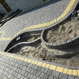 Układanie kostki brukowej na posesji, widoczne etapy prac ziemnych i przygotowania podłoża, obrzeża z kostki w kolorze grafitowym z żółtymi akcentami, częściowo zasypane piaskiem.