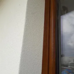 Fragment ocieplonej elewacji budynku z fakturą baranka, przylegającej do brązowej ramy okiennej z widocznym odblaskiem nieba i otoczenia.