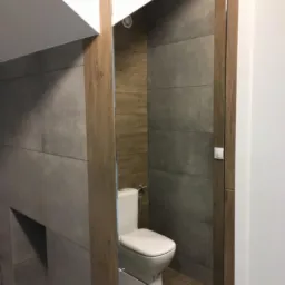 Nowoczesna łazienka z szarymi płytkami imitującymi beton, drewnianymi akcentami i białą toaletą, widoczna częściowo w lustrze, oświetlenie sufitowe punktowe.