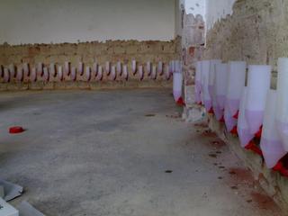 Ściana z widocznymi nawiertami i aplikatorami do iniekcji krystalicznej, częściowo zakryta plastikowymi pojemnikami z różowym płynem, przygotowana do wzmocnienia i osuszenia muru.