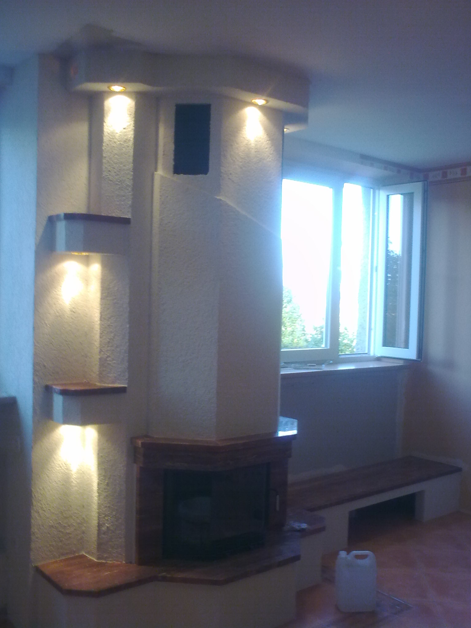 Murowany kominek z półkami oświetlonymi punktowymi lampami, wykończony marmurem, z siedziskiem wbudowanym w ścianę przy oknie.