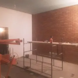 Ściana wyłożona cegłą klinkierową w trakcie prac, widoczne rusztowanie, mieszadło do zaprawy, narzędzia i butelka ze spryskiwaczem, surowy stan wykończenia.