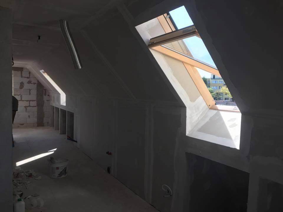 Poddasze w trakcie prac wykończeniowych: widoczne okno dachowe z drewnianą ramą, ściany pokryte płytami gipsowo-kartonowymi przed szpachlowaniem, fragment instalacji wentylacyjnej pod sufitem...