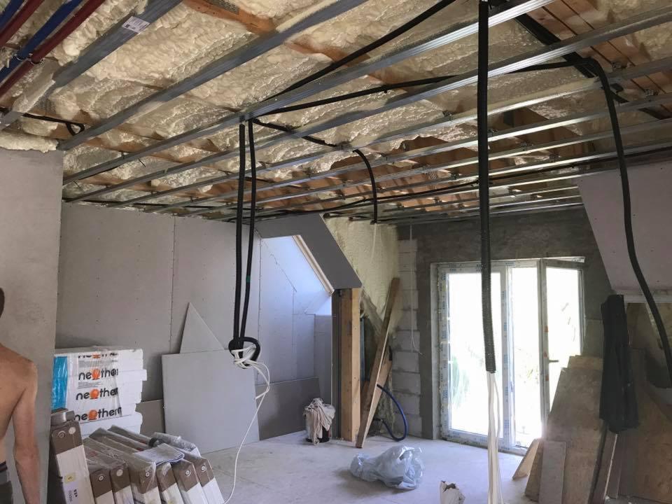 Wnętrze w trakcie prac wykończeniowych: sufit z izolacją piankową, metalowa konstrukcja podwieszana, widoczne rury instalacyjne, ściany z płyt gipsowo-kartonowych, okno balkonowe i materiały...