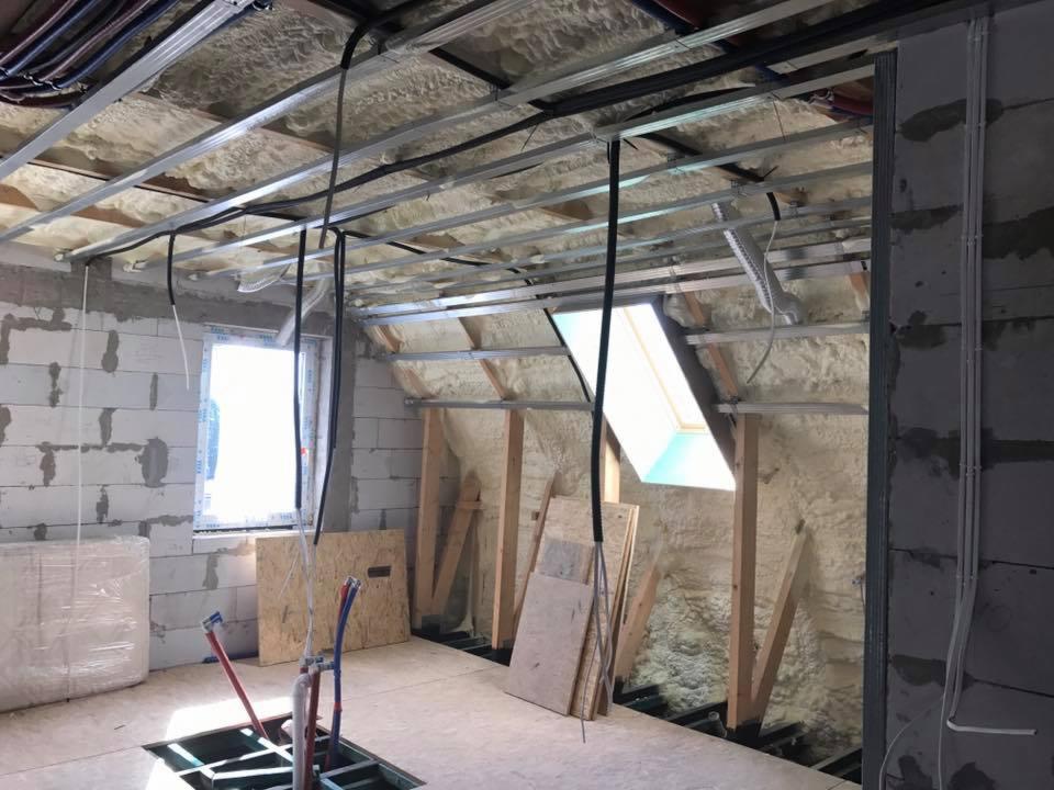 Poddasze w trakcie adaptacji: ocieplenie pianą PUR na skosach dachu, metalowa konstrukcja pod sufitem, instalacje wodne wystające z podłogi, okno dachowe wpuszczające jasne światło.