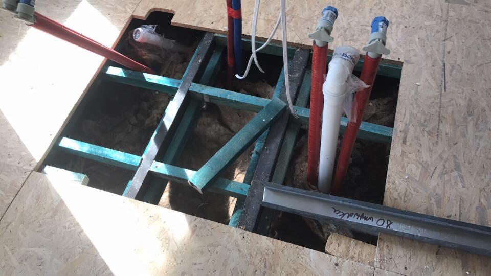 Otwór w podłodze z widocznymi rurami instalacji wodnej w kolorach czerwonym i niebieskim, oraz białą rurą PCV, z fragmentami drewnianej konstrukcji i izolacji w tle.