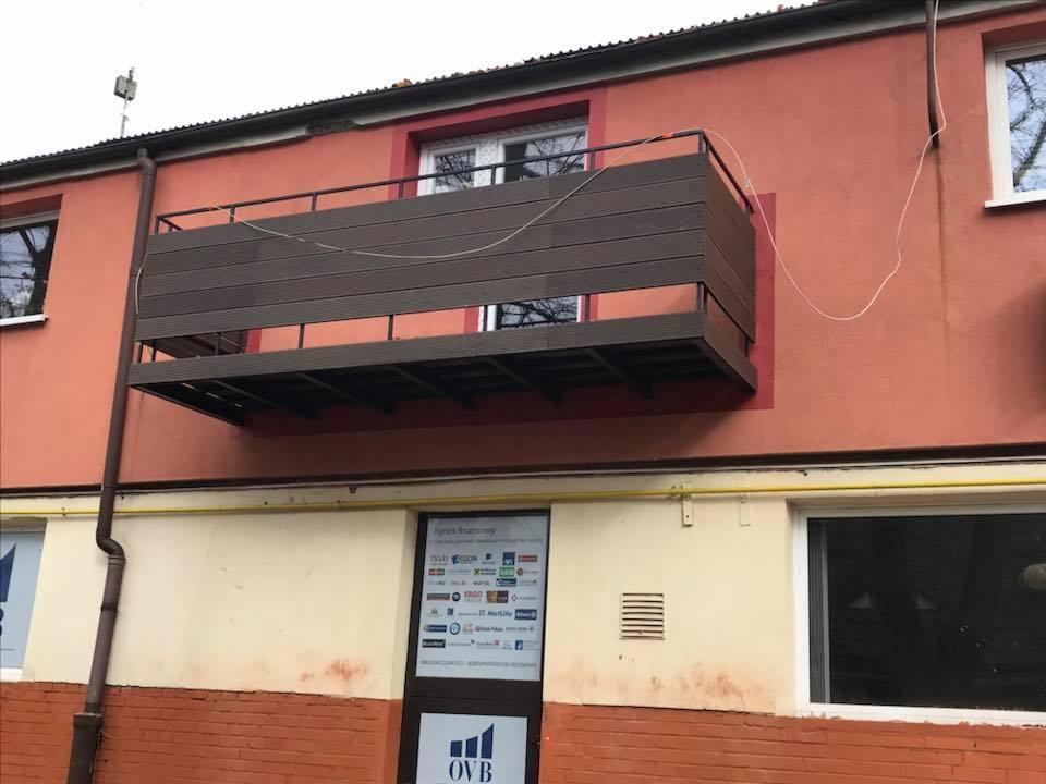 Zamontowany balkon z desek kompozytowych na czerwonym budynku, widoczny z zewnątrz, z rurą spustową po lewej stronie i oknem na górze.