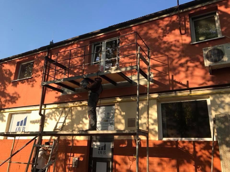 Pracownik na rusztowaniu spawa metalową konstrukcję balkonu przy dwupiętrowym budynku z pomarańczową elewacją, widoczne okna z żaluzjami i klimatyzator na ścianie.