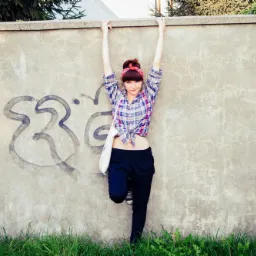 Młoda kobieta w kraciastej koszuli i czarnych spodniach, z czerwoną opaską na włosach, opiera się o betonowy mur z graffiti i zielonymi pnączami.