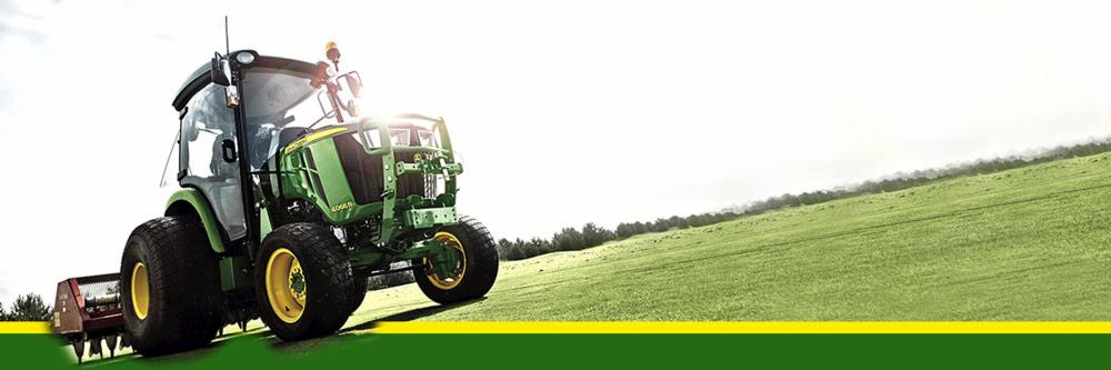 Zielony traktor John Deere 4066R na zielonym polu, z zamontowanym sprzętem rolniczym z tyłu, gotowy do pracy.