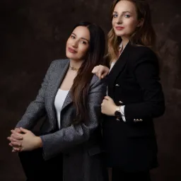 Portret dwóch kobiet w eleganckich marynarkach na tle ciemnego, teksturowanego tła. Kobieta po lewej w szarej marynarce siedzi z założonymi rękami, uśmiechając się. Kobieta po prawej, w czarnej...