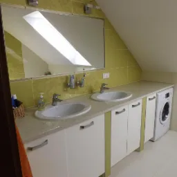 Ukończona łazienka na poddaszu ze skośnym sufitem: dwie białe umywalki z szafkami, pralka i lustro odbijające okno dachowe. Żółte płytki z motywem roślinnym.