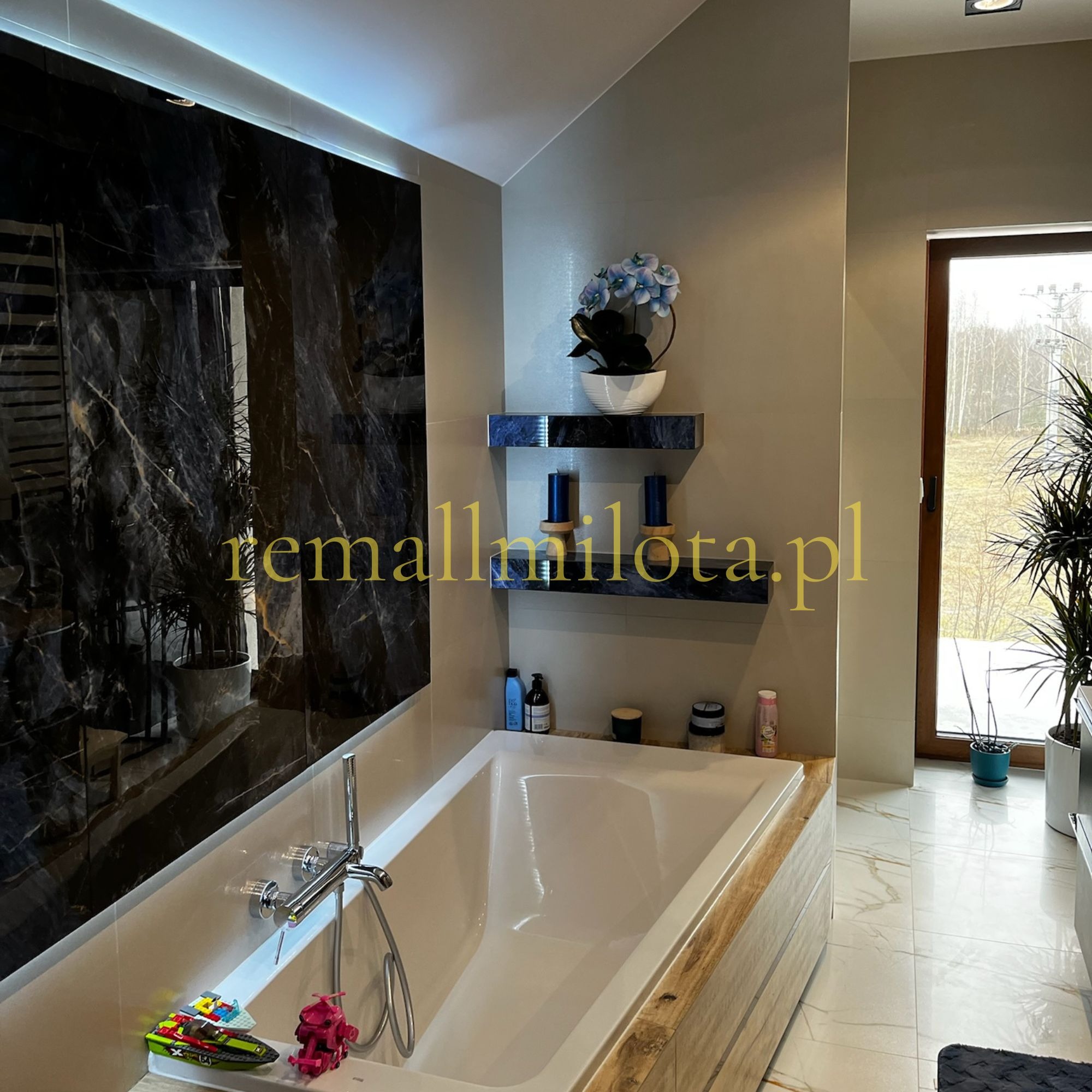 Nowoczesna łazienka z wanną obudowaną drewnem, czarnym marmurem na ścianie i widokiem na zewnątrz. Na wannie leżą dziecięce zabawki, dodając wnętrzu domowego charakteru.