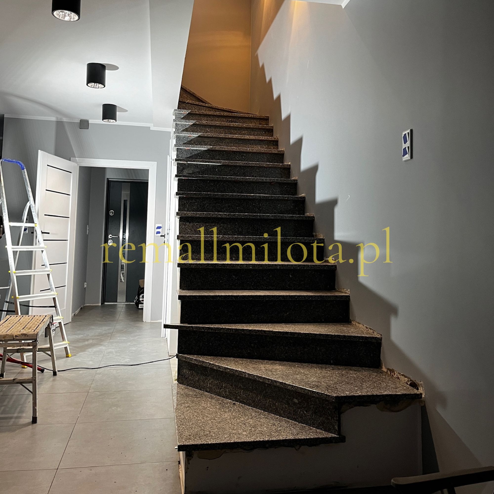 Granitowe schody spiralne w szarej klatce schodowej, widoczne niedokończone wykończenia, drabina i stołek malarski w tle. Oświetlenie sufitowe.