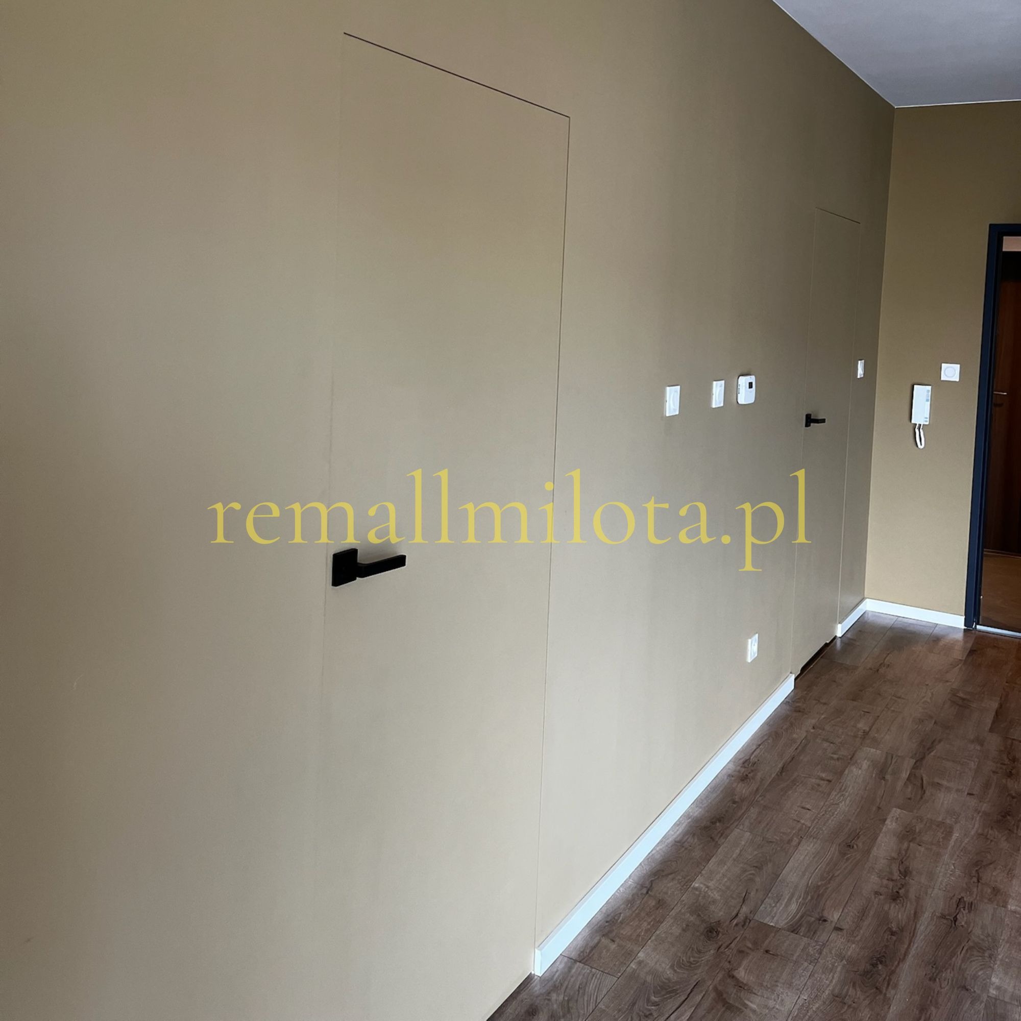 Beżowa ściana z ukrytymi drzwiami i czarną klamką. Widoczne listwy przypodłogowe i panele podłogowe. Minimalistyczny design wnętrza z elementami wykończeniowymi.