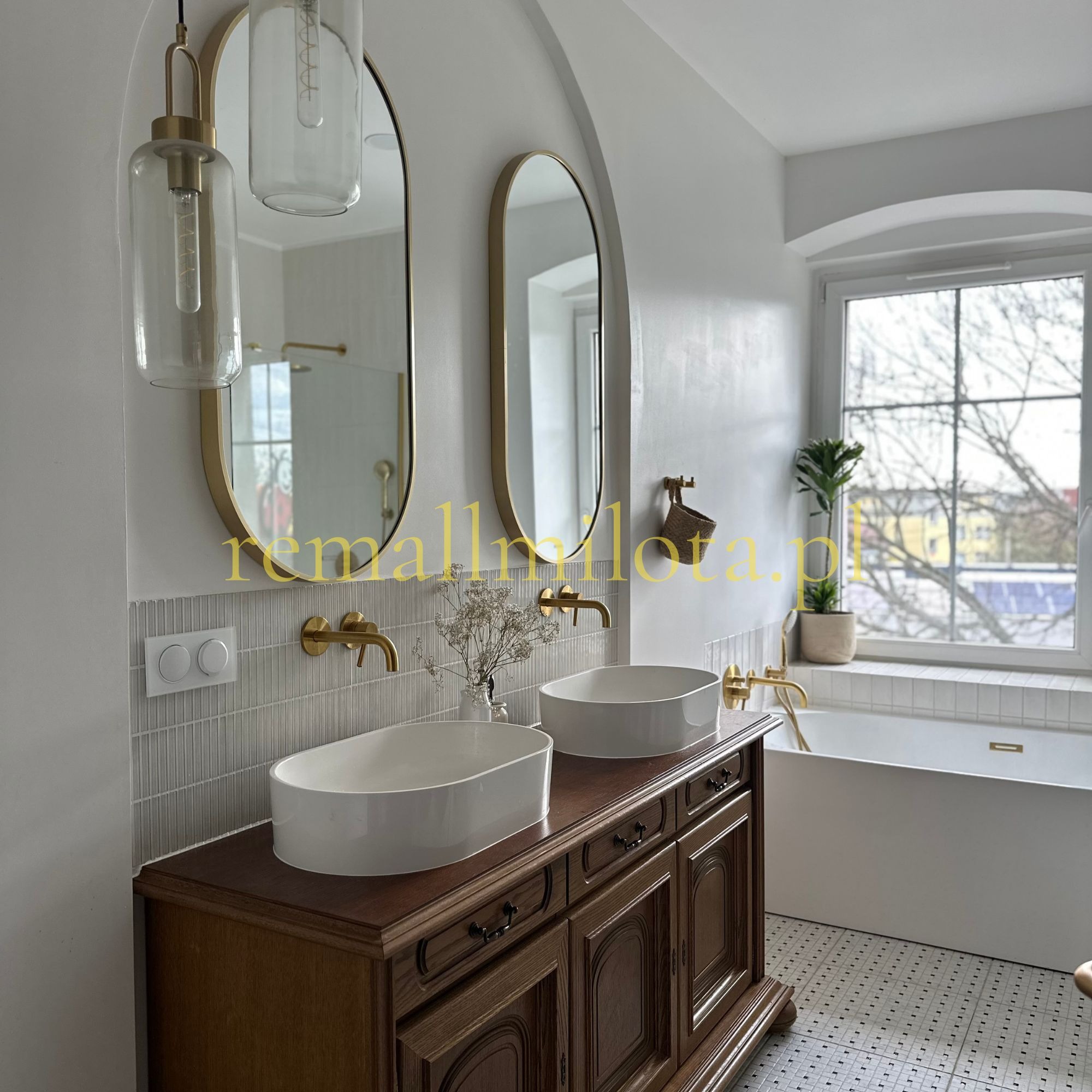 Stylowa łazienka z drewnianą szafką pod umywalki, owalnymi lustrami w złotych ramach i wiszącymi lampami. Białe płytki i wanna przy oknie z widokiem na drzewa.