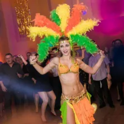 Uśmiechnięta kobieta w barwnym kostiumie z piór, tańcząca na tle bawiących się ludzi i dekoracji z frędzli na imprezie.