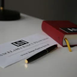 Wizytówka kancelarii prawnej, czarny długopis i czerwona książka z zakładką na białym biurku.