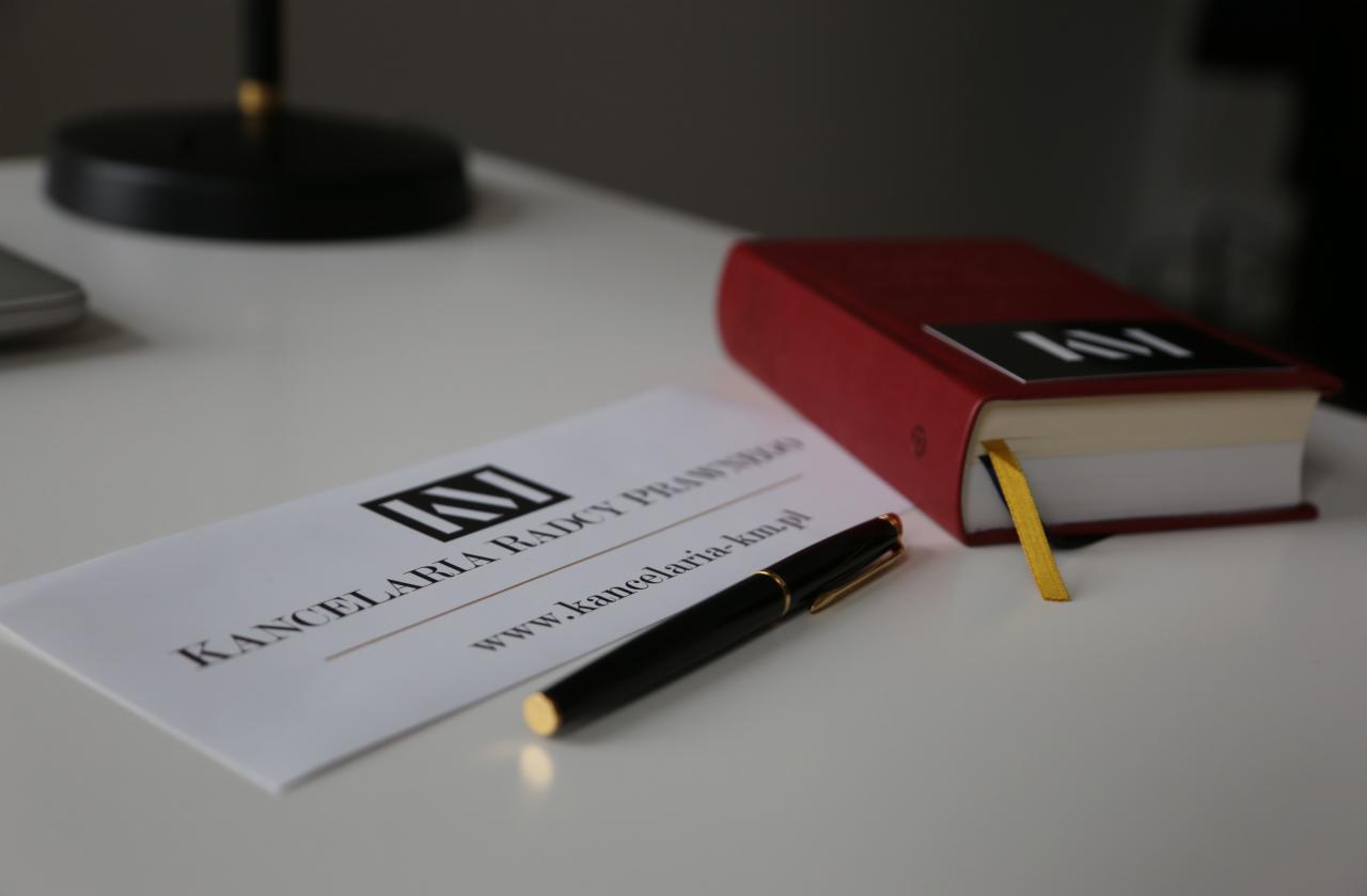 Wizytówka kancelarii prawnej, czarny długopis i czerwona książka z zakładką na białym biurku.