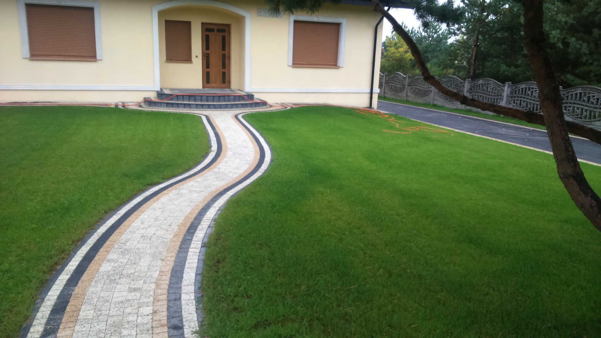 Kręty chodnik z jasnej kostki brukowej z czarnymi i beżowymi paskami obramowania prowadzi od schodów wejściowych do domu z kremową elewacją przez idealnie przystrzyżony trawnik.