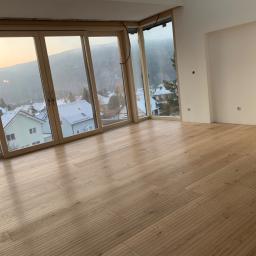 Firma Chrapla - Jasny, drewniany parkiet ułożony w przestronnym wnętrzu z dużymi oknami, za którymi widać ośnieżone dachy domów i zalesione wzgórza o zmierzchu.