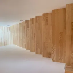 Nowoczesne wnętrze z drewnianą ścianą z paneli ułożonych pionowo, jasna podłoga i okno z widokiem na zewnątrz.