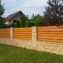 Nowoczesne ogrodzenie z poziomych desek w ciepłym odcieniu drewna, osadzone na niskim murku z kamienia piaskowego, z budynkiem w tle.