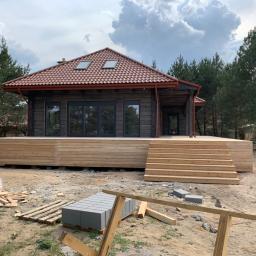 Firma Chrapla - Drewniany dom z ciemną elewacją i jasnym tarasem w trakcie budowy, widoczne drewniane schody i porozrzucane materiały budowlane.