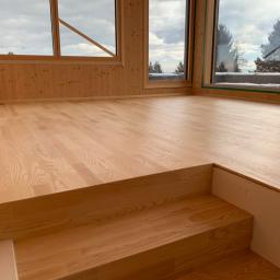 Firma Chrapla - Jasny, drewniany parkiet ułożony w nowym domu z drewnianymi ścianami i dużymi oknami z widokiem na zimowy krajobraz.