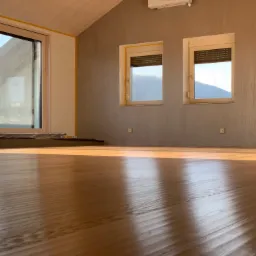 Jasne, nowo wycyklinowane podłogi w pokoju z dwoma oknami z roletami i dużym oknem przesuwnym, widok na góry w tle.