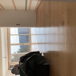 Firma Chrapla - Jasne drewniane deski podłogowe, widok na okno z krajobrazem górskim, częściowo zasłonięte workiem budowlanym i pudełkami, listwy sufitowe w trakcie montażu.