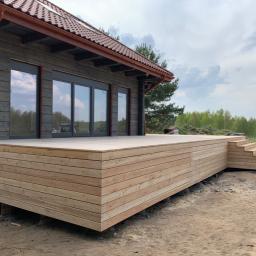 Firma Chrapla - Jasna drewniana platforma tarasowa z schodami, przylegająca do ciemnego drewnianego domu z brązowym dachem, widok z boku na tle zieleni i pochmurnego nieba.