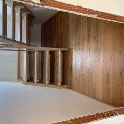 Firma Chrapla - Widok z góry na drewniane schody w trakcie montażu, z widocznymi zabezpieczeniami na stopniach i nową drewnianą podłogą, częściowo wykończone ściany z widocznymi fragmentami cegieł.