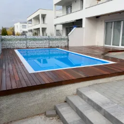 Nowoczesny basen otoczony ciemnym drewnianym tarasem, z gabionami w tle i minimalistyczną architekturą domu.