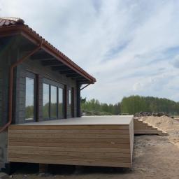 Firma Chrapla - Nowoczesny dom z ciemnego drewna z przylegającym tarasem z jasnego drewna, schody prowadzące na taras, w tle las i plac budowy.