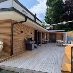 Firma Chrapla - Nowoczesny dom z drewnianą elewacją i tarasem z jasnego drewna, na którym stoi grill, krzesło wiszące i stół z krzesłami. Widoczna osoba pracująca na tarasie.