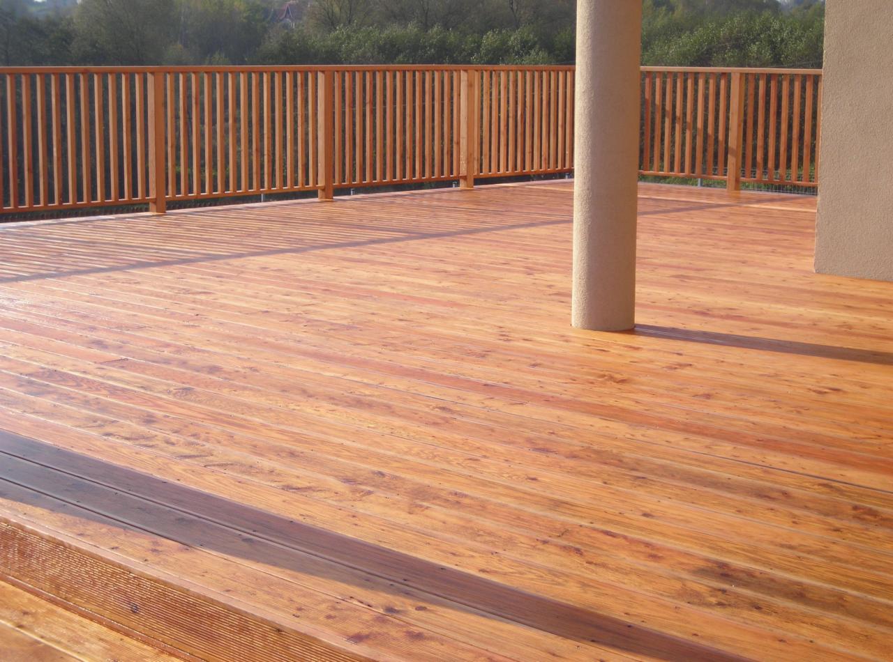 Nowoczesny taras z desek w odcieniach brązu, otoczony drewnianą balustradą, z widocznym słupem nośnym i fragmentem ściany budynku.