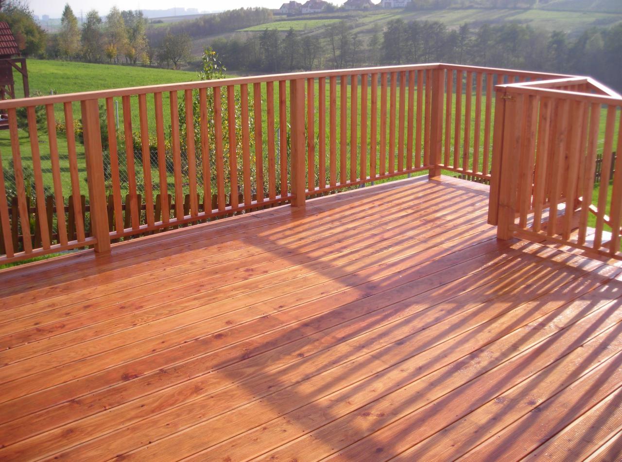 Świeżo polakierowany drewniany taras z balustradą, widok na zielone wzgórza w oddali, długie cienie rzucane przez popołudniowe słońce.