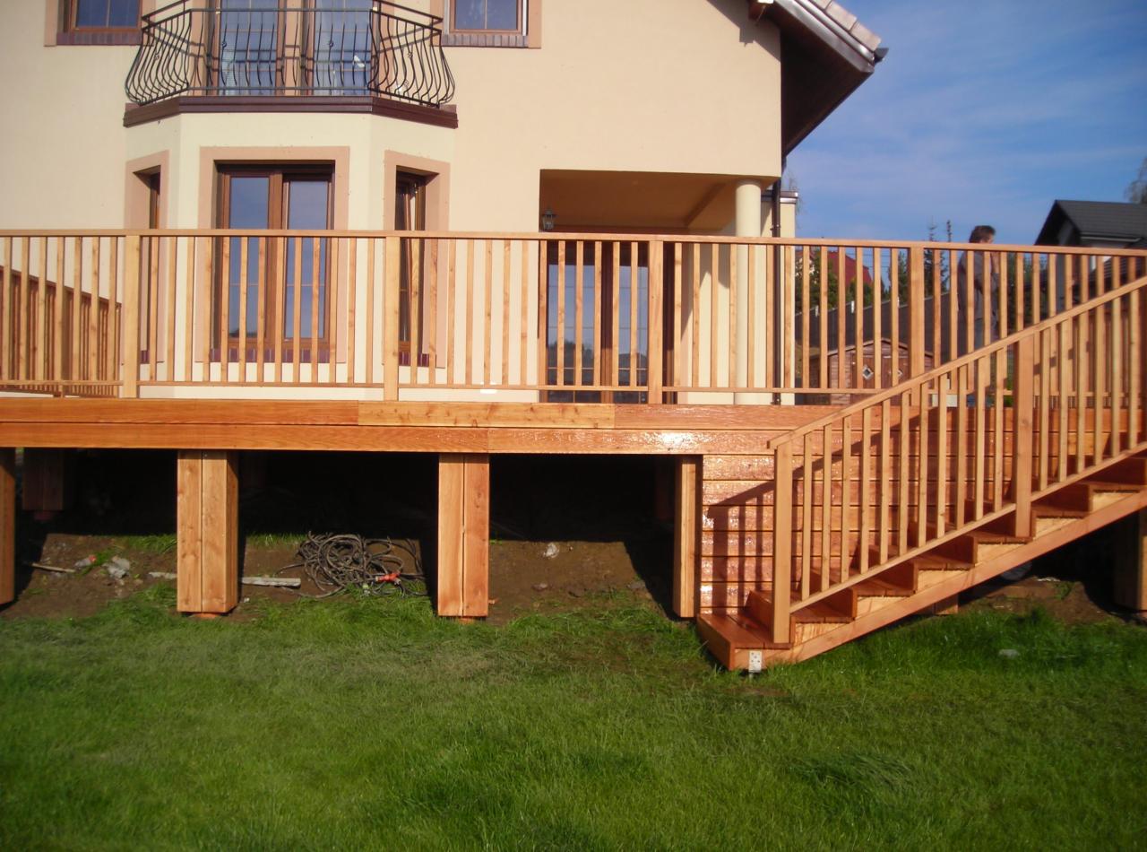 Świeżo zaimpregnowany taras z jasnego drewna z balustradą i schodami, widok z poziomu trawnika, na tle budynku z balkonem.