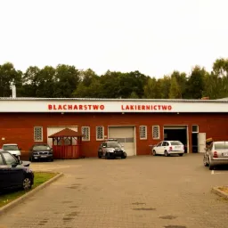 Budynek warsztatu z czerwonej cegły z napisem na górze. Na placu przed budynkiem zaparkowane samochody.