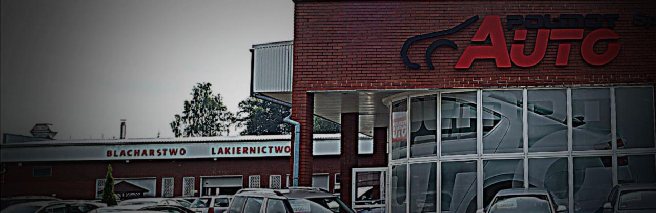 Fasada budynku warsztatu samochodowego z napisem 'Blacharstwo Lakiernictwo' oraz logo 'AUTO' na ceglanej ścianie, widoczne samochody na parkingu.