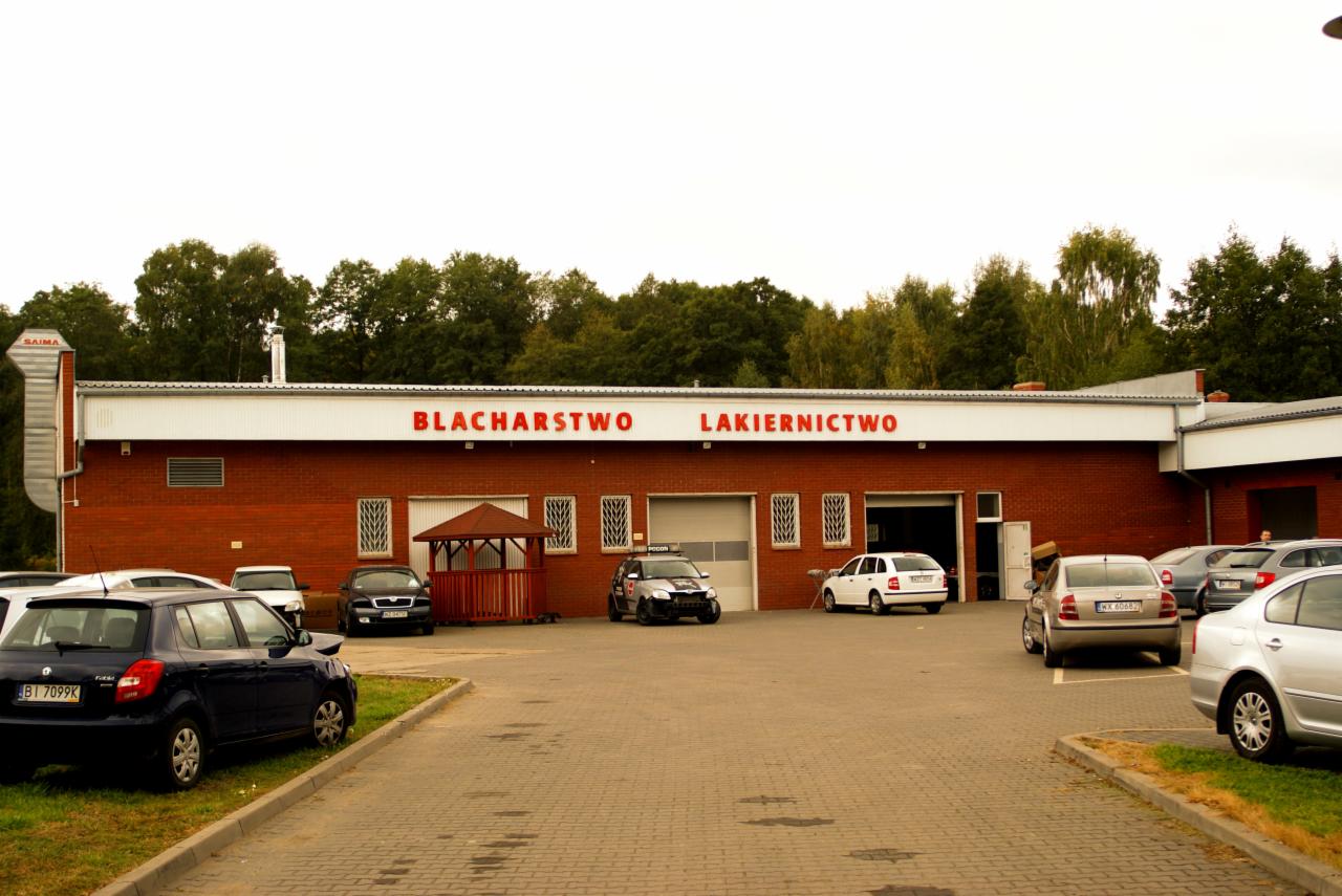 Budynek warsztatu z czerwonej cegły z napisem na górze. Na placu przed budynkiem zaparkowane samochody.