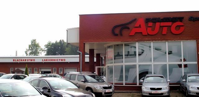 Budynek warsztatu samochodowego z szyldem 'AUTO' i napisem 'Blacharstwo Lakiernictwo', przed budynkiem zaparkowane samochody.