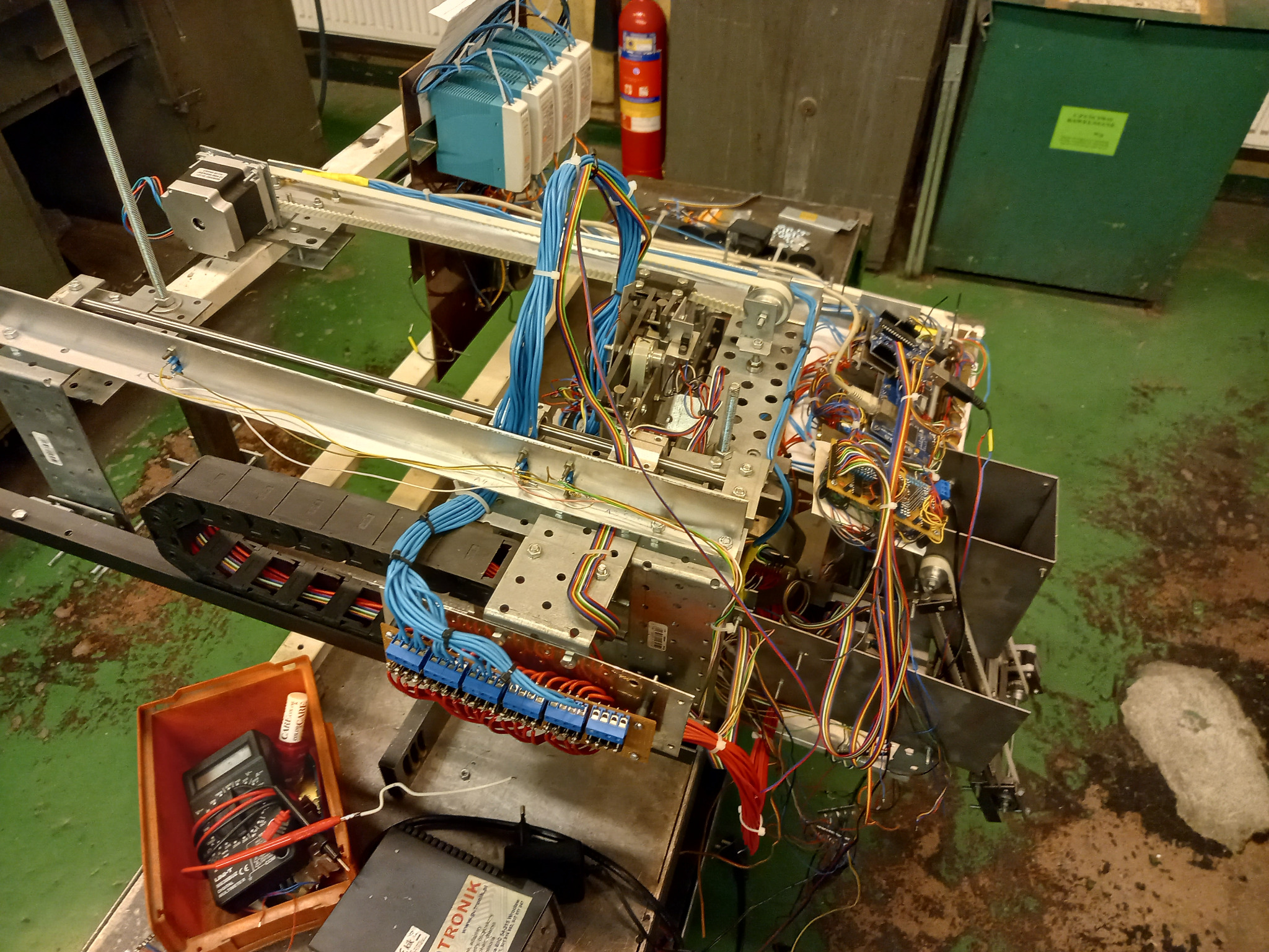 Widok z góry na metalową konstrukcję z licznymi przewodami, złączkami i elementami elektronicznymi, w tle zielona podłoga i gaśnica, w pomarańczowym pojemniku leży multimetr.