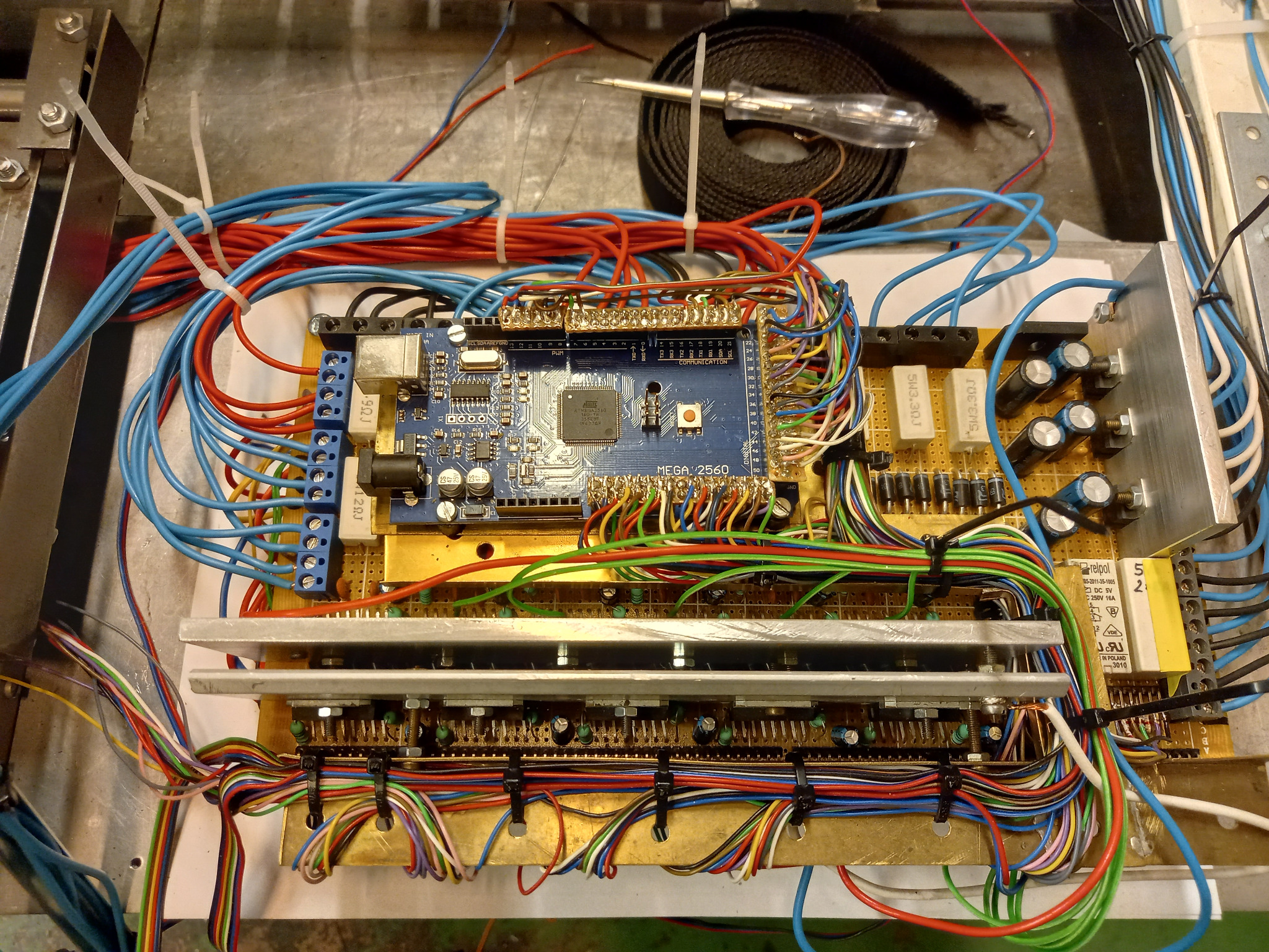 Panel elektryczny z płytką Arduino Mega 2560, licznymi przewodami w różnych kolorach, kondensatorami i rezystorami, w trakcie montażu lub testowania, widoczny tester napięcia.