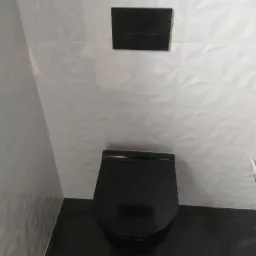 Czarna toaleta podwieszana z czarnym przyciskiem spłukującym na ścianie wyłożonej białymi, trójwymiarowymi płytkami.