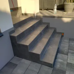 Wykończone schody wejściowe z szarych płytek ceramicznych, prowadzące na taras z widoczną butelką wody i donicą z rośliną w tle.