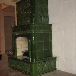 Kaflowy piec kominkowy w odcieniach zieleni, z dekoracyjnymi kaflami i paleniskiem z rusztem, ustawiony na brązowej podłodze z płytek, na tle surowej, białej ściany z widocznymi szkicami.